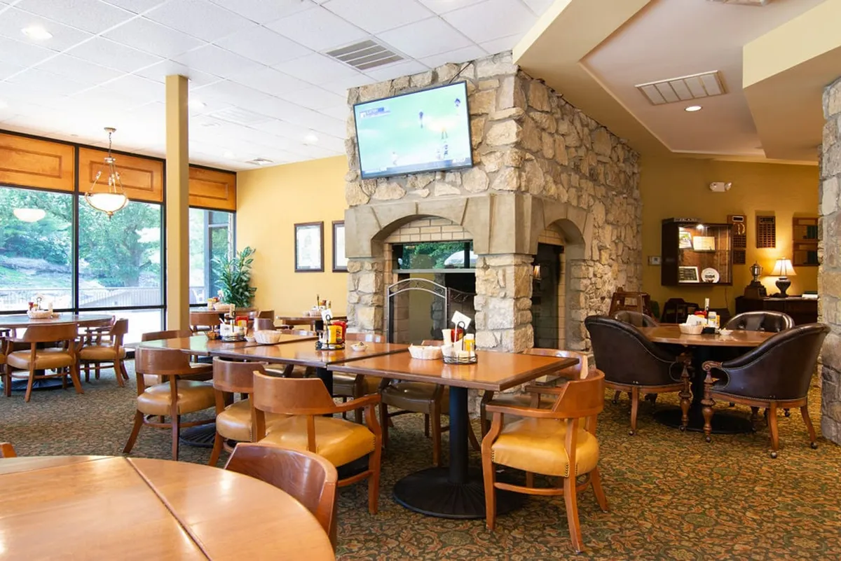 This interior image features a casually elegant clubhouse dining area with a stone fireplace, tables and chairs, a television screen for watching sports, and large windows offering views outside. The inviting atmosphere suits post-golf relaxation and gatherings with friends.