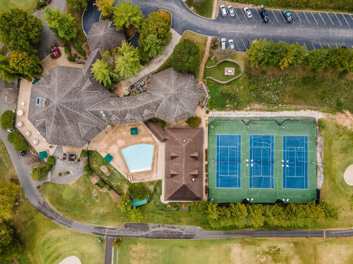 A bright exterior view featuring a large swimming pool with lounge chairs, adjacent tennis courts, and the main community building. The area is bustling with activity, inviting for both play and relaxation, and surrounded by lush greenery.
