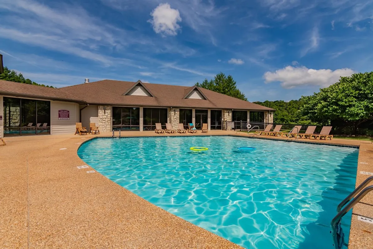 A sunlit swimming pool is accented by lounge chairs, with a modern building featuring large windows and green trees in the background. The cheerful outdoor setting encourages relaxation and socializing.
