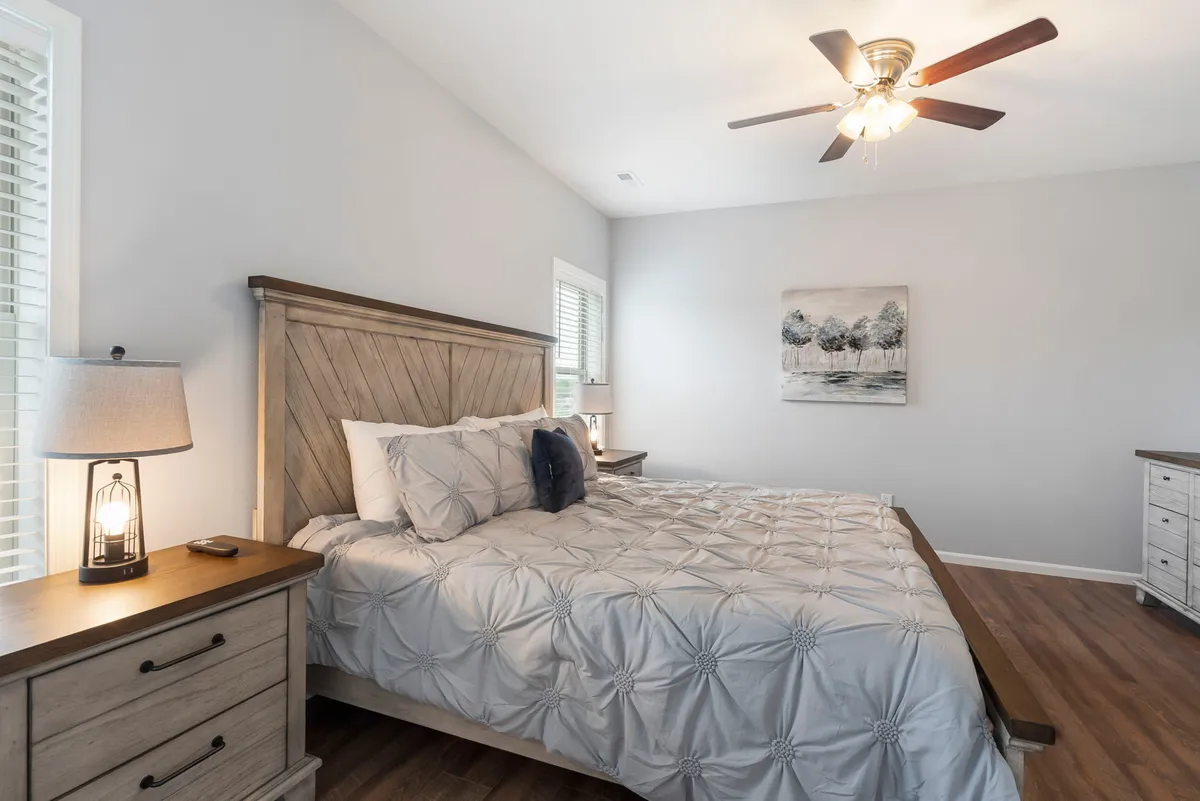 A tranquil bedroom scene featuring a neatly made bed with crisp linens, a nightstand adorned with a lamp, and tasteful artwork on the wall. Soft, natural light filters through a window, enhancing the serene and restful atmosphere.