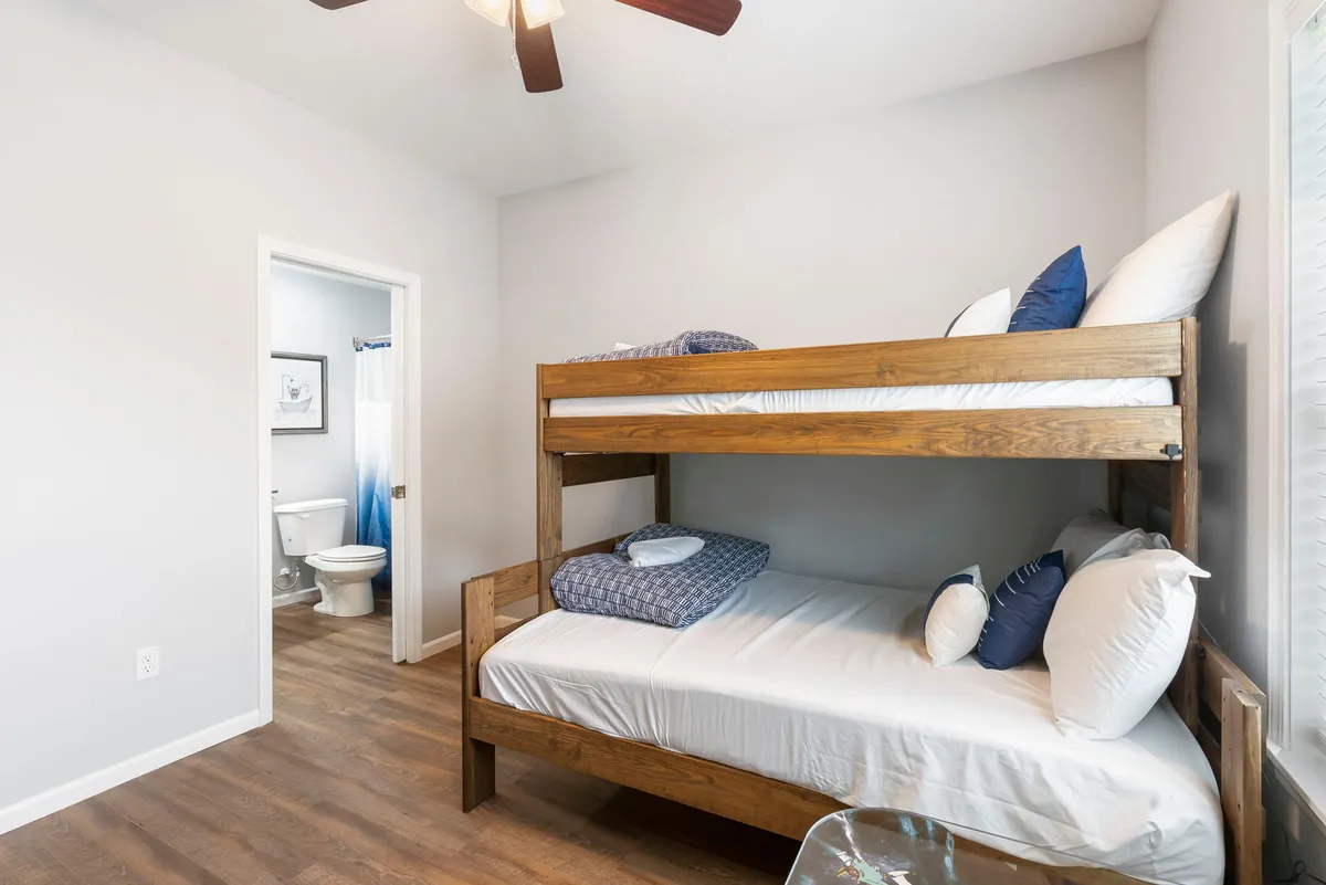A cozy third bedroom with a twin-over-full bunk bed dressed in decorative pillows. Natural light streams in from a window, and a doorway leads to a nearby bathroom, making the space ideal for children or families.