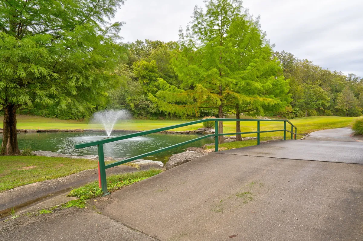 A picturesque bridge with green railings spans over calm water, with a fountain in the center and lush trees all around. The scene conveys tranquility and offers glimpses of seasonal foliage, making it a perfect spot for walking, birdwatching, and enjoying nature.