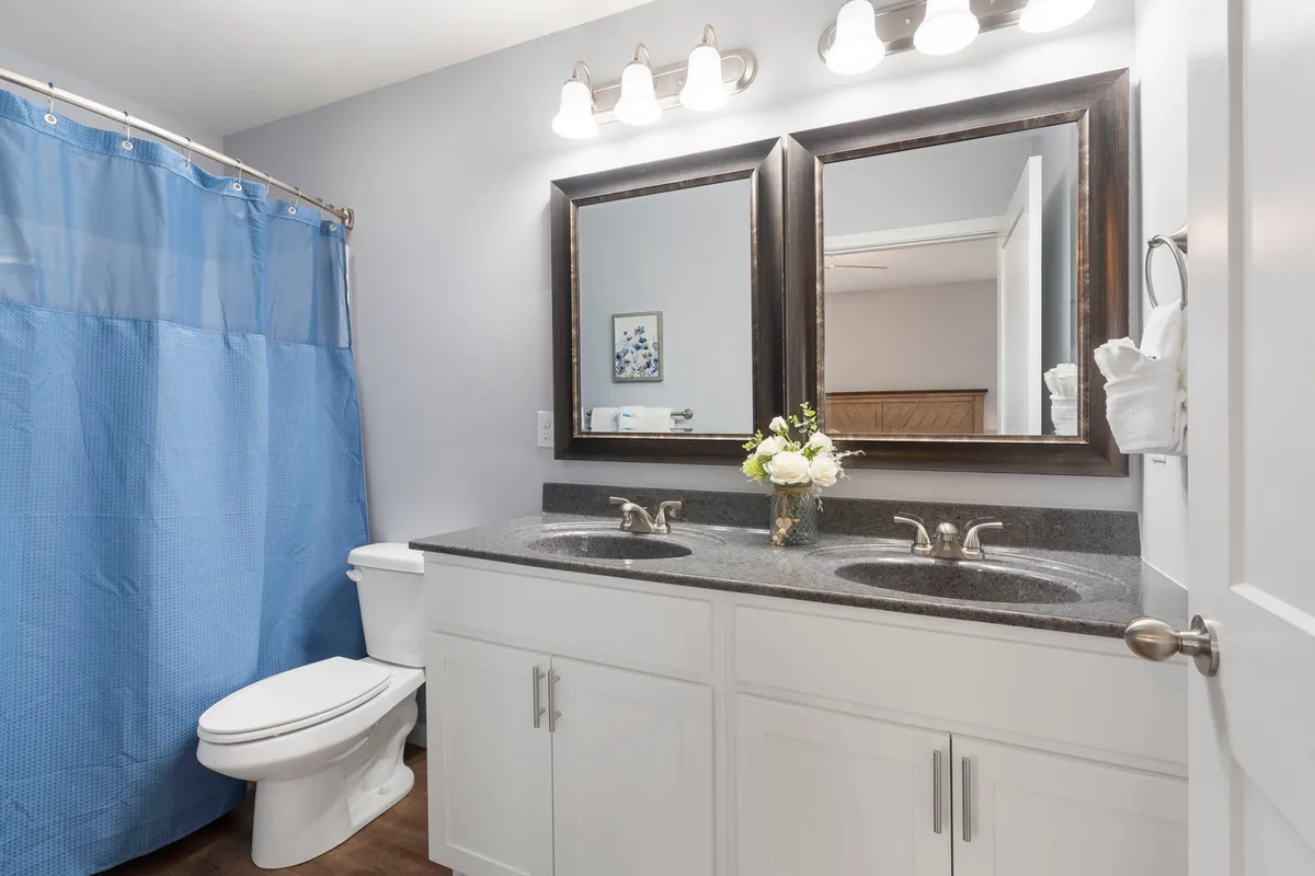 A modern bathroom offering a calming palette, double vanity sinks, ample mirrors, and a blue shower curtain. A floral arrangement adds a touch of luxury and freshness, suggesting a space for relaxation and rejuvenation.