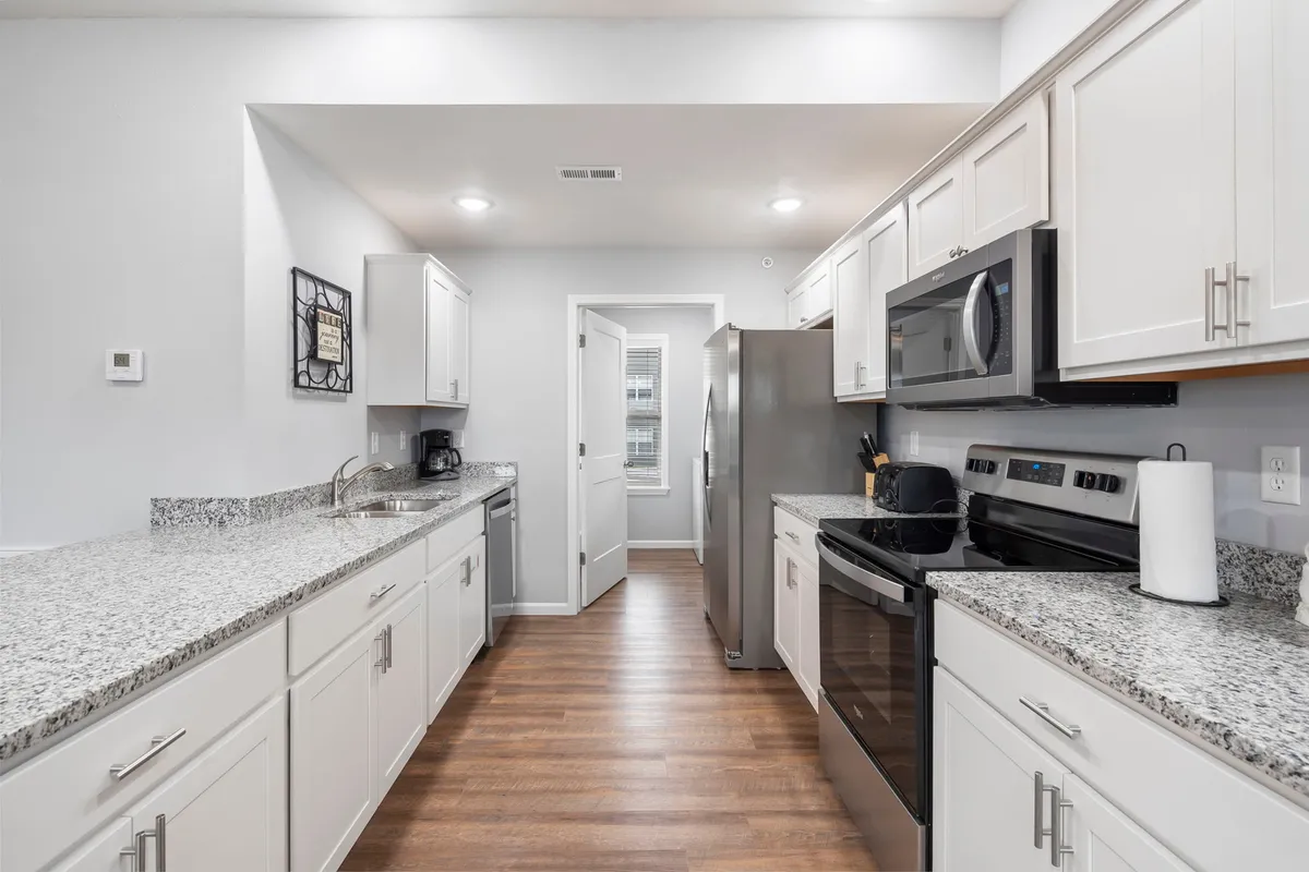 The image presents a modern, fully-equipped kitchen with granite countertops, stainless steel appliances, and white cabinetry. An open view into an adjacent room hints at the kitchen's integration with the broader living space, suggesting easy movement and interaction during gatherings.